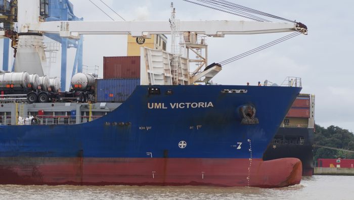 Navio cargueiro azul “UML VICTORIA” em manobra no porto, com vista do casco, proa e equipamentos de carga ao fundo.
