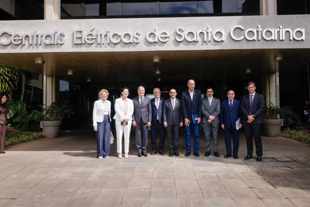 Grupo de pessoas posando em frente à fachada com a placa “Centrais Elétricas de Santa Catarina”, em foto institucional ao ar livre com arquitetura moderna ao fundo.