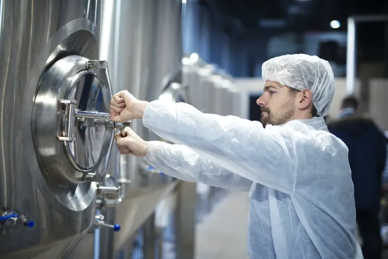 Trabalhador em laboratório com touca e roupa de proteção inspecionando uma máquina de aço inox em ambiente de fábrica, próximo ao painel e válvulas.