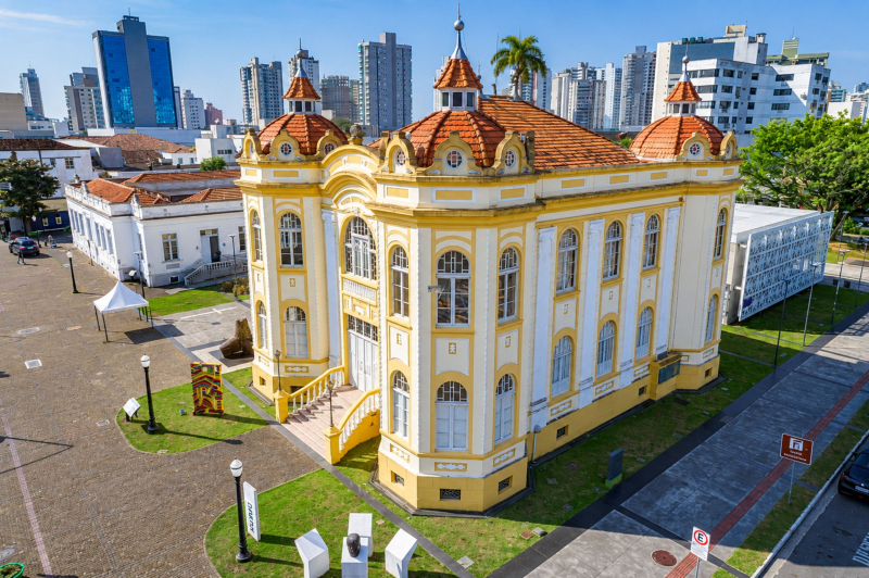 Vista aérea de prédio histórico amarelo e branco com telhado colonial e cúpulas, em um centro urbano com jardins, faixas de pedestres e prédios ao fundo.