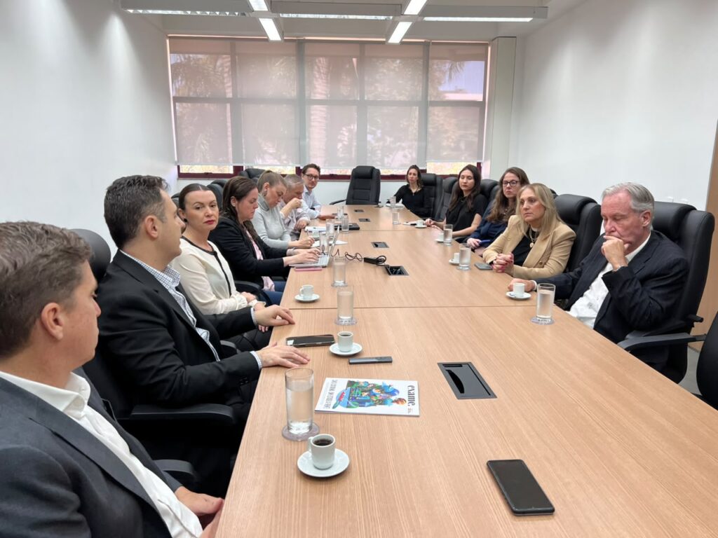 Reunião corporativa em sala de conferências com pessoas sentadas em cadeiras estofadas ao redor de uma mesa de madeira, com copos e celulares sobre a superfície