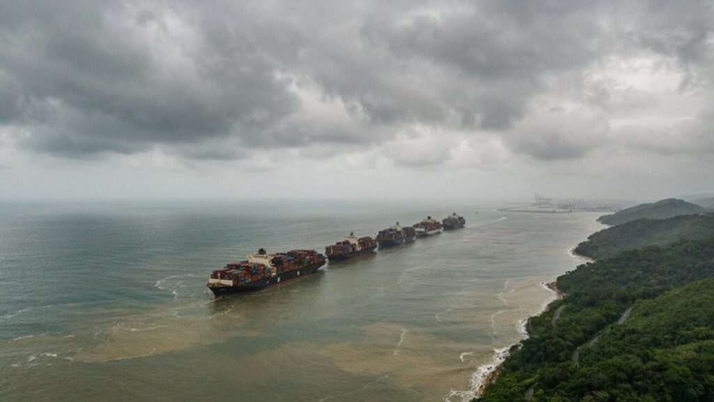 Vista aérea de navios cargueiros atracados ao longo da costa, com o mar ao fundo e uma linha de floresta verde à direita sob céu nublado, ao entardecer