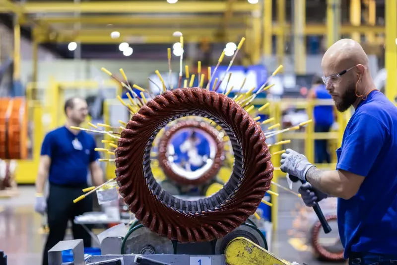 Trabalhador industrial em linha de produção utilizando uma ferramenta elétrica para polir um disco circular de metal marrom, em ambiente de fábrica com máquinas ao fundo.