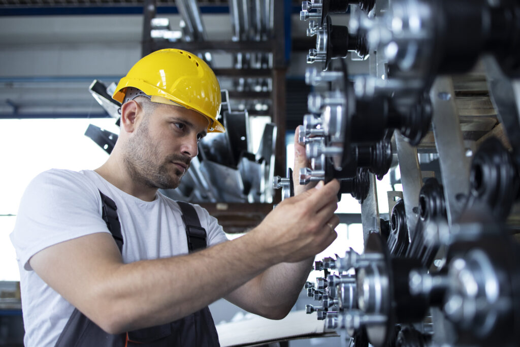 Trabalhador industrial com capacete amarelo e uniforme ajustando componentes mecânicos em uma máquina durante manutenção na fábrica, com foco na operação e segurança.