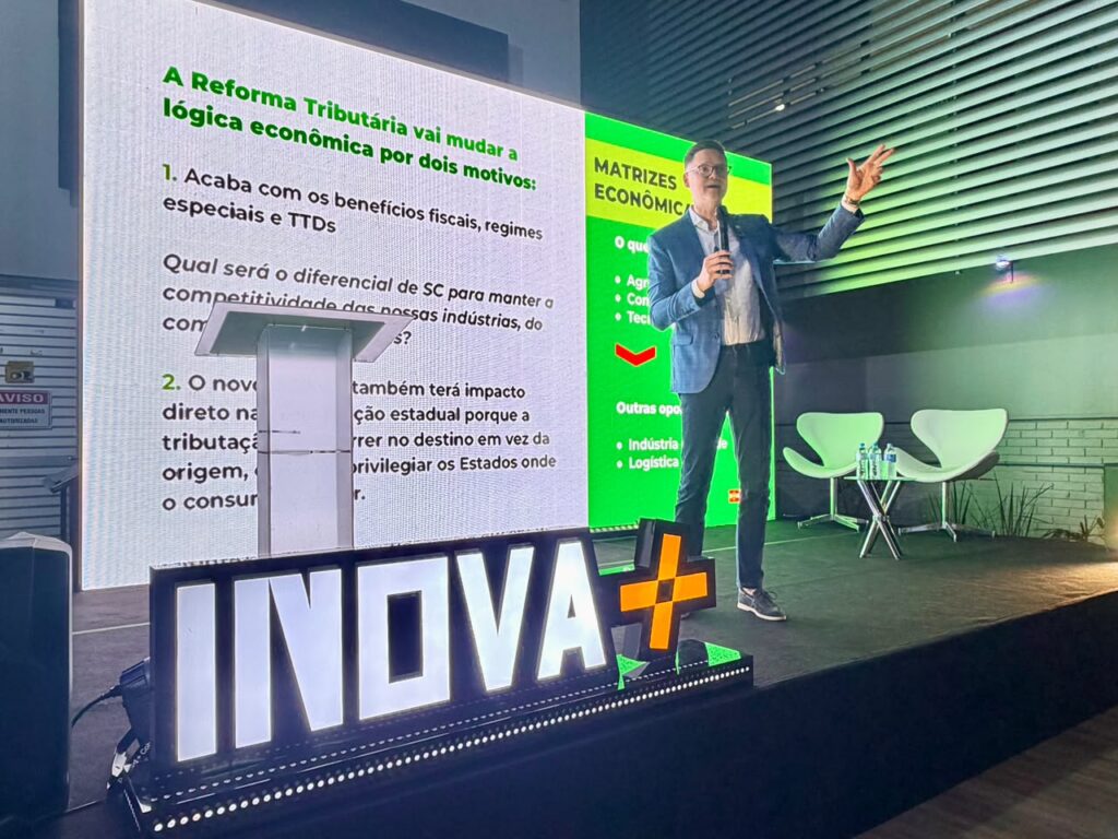 Palestra em evento corporativo com apresentador em pé ao lado de um telão que mostra texto sobre “A Reforma Tributária” e “MATRIZES ECONÔMICAS”, com destaque para gráficos e tópicos.