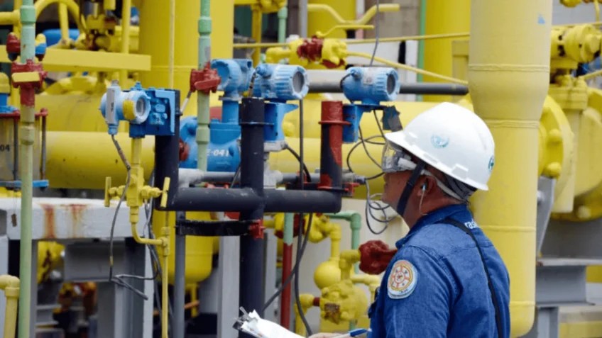 Trabalhador com capacete de proteção em área industrial com tubulações amarelas e azuis, válvulas e equipamentos de processo ao fundo.