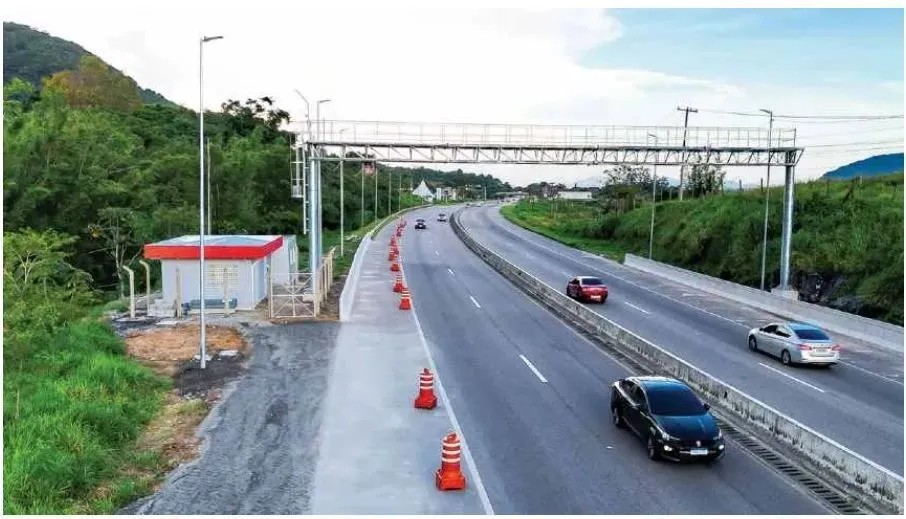Trecho de rodovia em obra com cones de sinalização, tapume e equipes ao lado da pista, incluindo veículos trafegando sob uma estrutura metálica em área verde ao fundo.