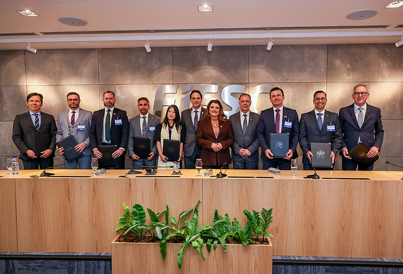 Grupo de autoridades e participantes em uma mesa de conferência, em evento corporativo, com placas e credenciais ao fundo e plantas ornamentais no centro.