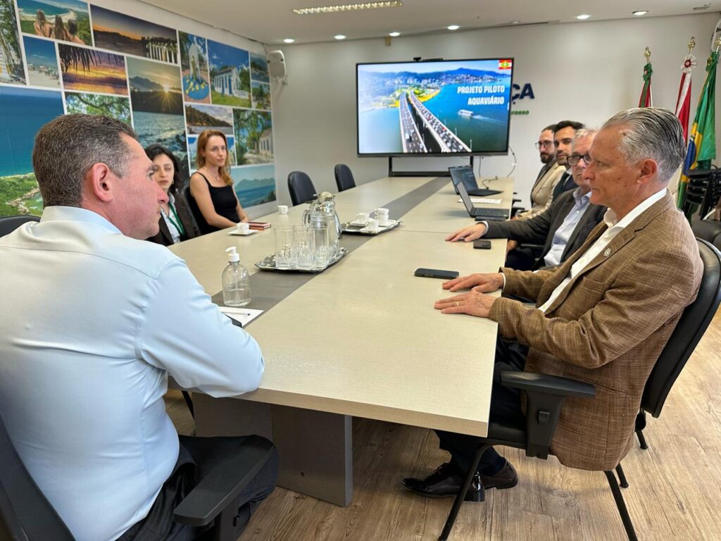 Reunião em sala de comitê com participantes ao redor de uma mesa, enquanto um monitor exibe a apresentação “PROJETO PILOTO” e “AQUIDAUANA”.