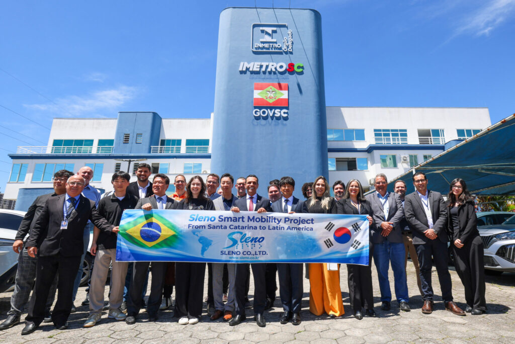 Grupo de pessoas em evento corporativo em frente ao prédio do IMETROSC, com faixa de apresentação “Sleno Smart Mobility Project” e bandeiras de Brasil, Santa Catarina e Coreia do Sul ao fundo do letreiro GovSC.