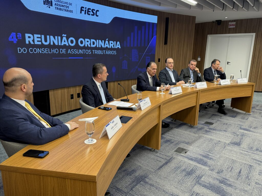 Quatro participantes em reunião em sala de conferências, com mesa de madeira e documentos, falando em microfones diante de telão com os dizeres “4ª REUNIÃO ORDINÁRIA DO CONSELHO DE ASSUNTOS TRIBUTÁRIOS” e logotipo FIESC.
