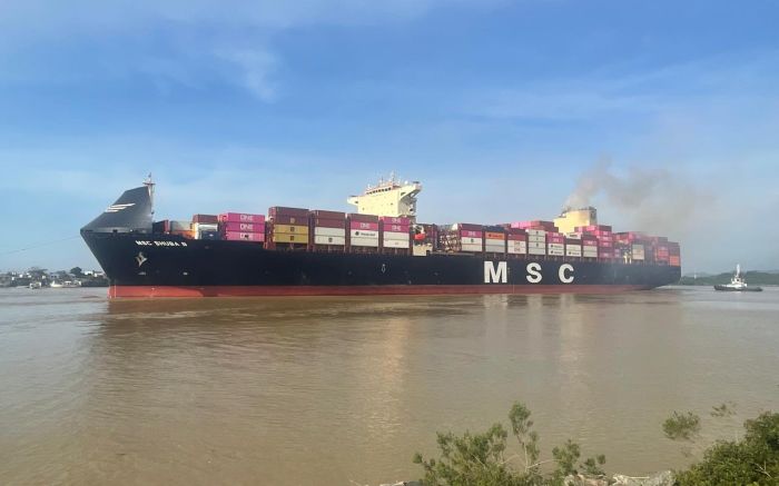 Navio porta-contêineres MSC carregado com caixas em um rio, visto à distância ao pôr do sol, com céu azul claro e água refletindo a embarcação
