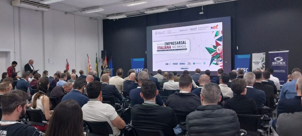 Palestra em auditório com plateia sentada assistindo apresentação em telão, em evento empresarial com telões de patrocinadores e bandeiras ao fundo.