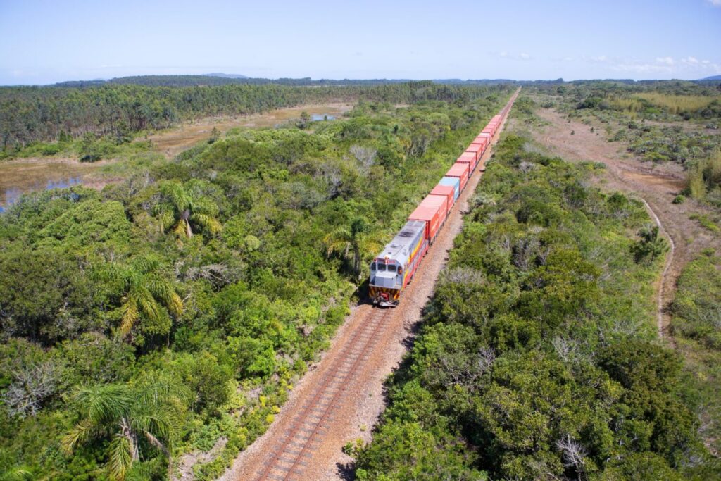 Vista aérea de um trem de passageiros atravessando uma área de vegetação densa, com vagões coloridos em uma linha férrea ao longo de trilhas de terra em meio ao cerrado e morros ao fundo.