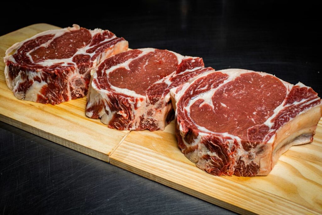 Cortes de carne bovina com marmoreio em fatias, sobre uma tábua de madeira, com aparência suculenta e gordura branca evidente, fotografados em fundo escuro.