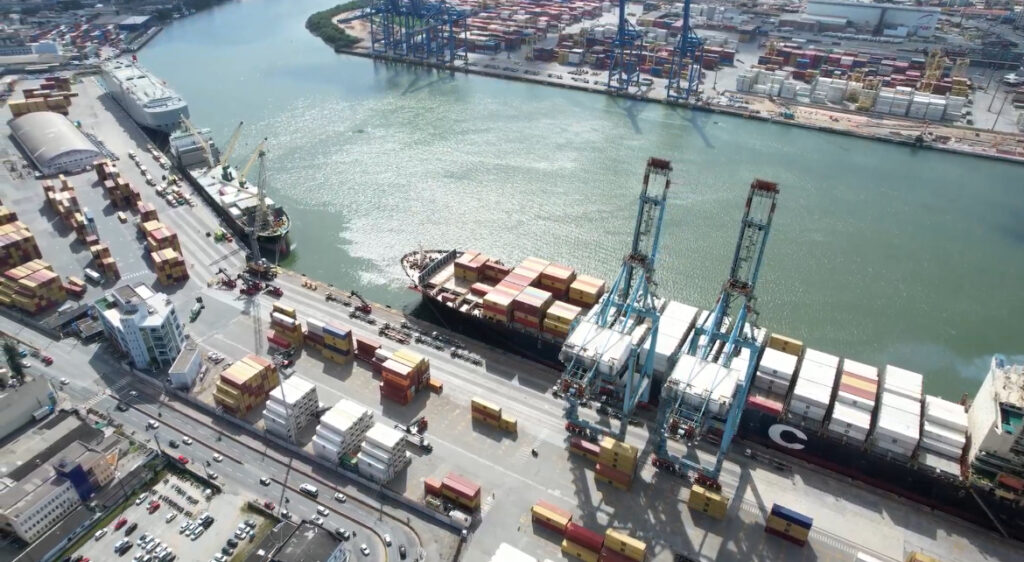 Vista aérea de um porto com navios cargueiros, contêineres empilhados e guindastes no cais, com tráfego de caminhões na área portuária.