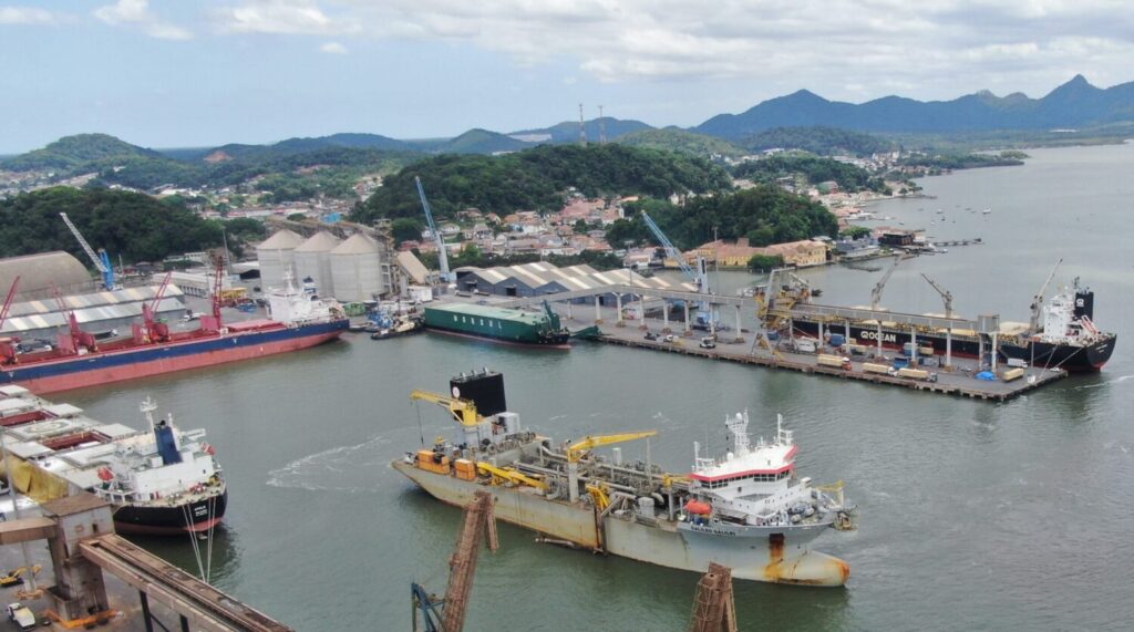 Vista aérea de um porto com navios atracados, guindastes e estruturas industriais ao lado de montanhas e da costa ao fundo.