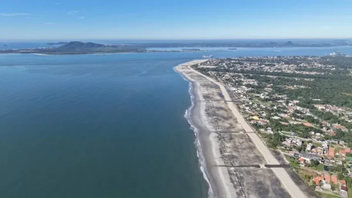 Vista aérea de uma praia extensa com faixa de areia, mar calmo e cidade à direita, mostrando ruas próximas à orla e vegetação ao fundo