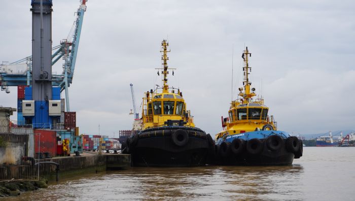 Dois rebocadores marítimos amarelos e pretos posicionados ao lado de um cais, com um navio cargueiro ao fundo e guindastes no porto.