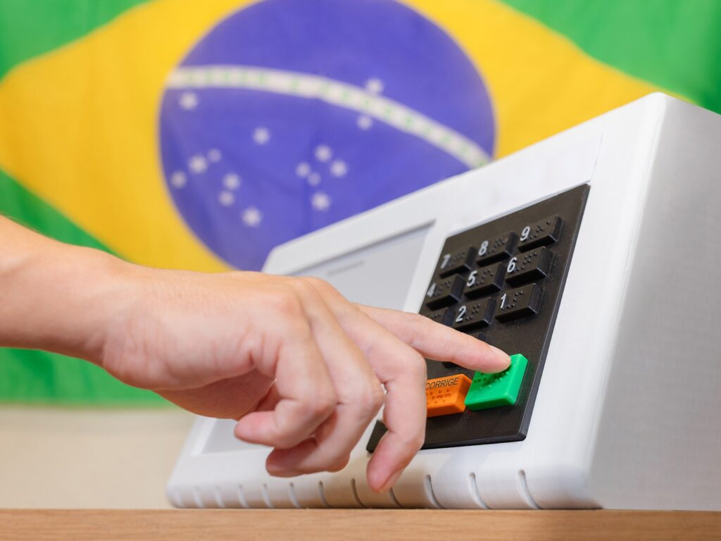Pessoa acionando um painel de seleção com teclas numéricas e luzes, em um brinquedo interativo sobre mesa de madeira; ao fundo há estampa colorida com arco-íris e estrelas.