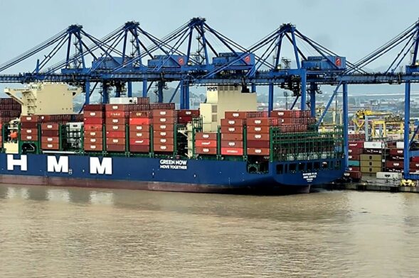 Navio porta-contêineres azul no porto, com guindastes industriais ao fundo e vários contêineres empilhados no convés, visto à beira da água.