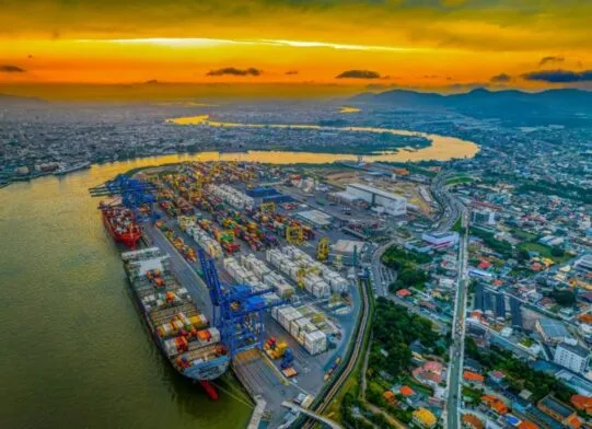Vista aérea do porto com navios e contêineres ao longo do rio, com ruas e áreas urbanas ao fundo sob um céu com nuvens e pôr do sol.