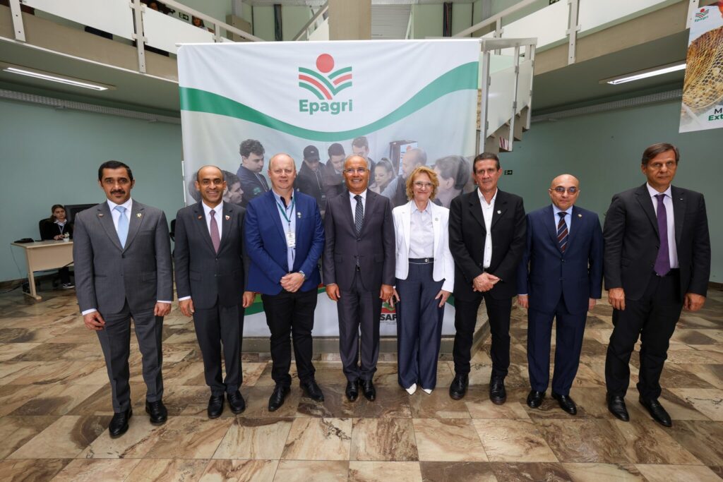 Grupo de autoridades em foto oficial em evento, em pé ao lado de uma grande bandeira com o logo da Epagrí, em ambiente institucional com piso de mármore.