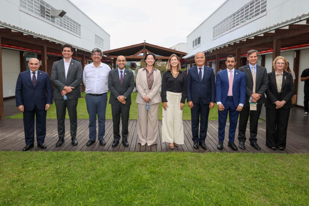 Foto em ambiente externo corporativo com um grupo de pessoas usando ternos e vestidos, posando em um jardim com gramado e área de madeira entre dois prédios.
