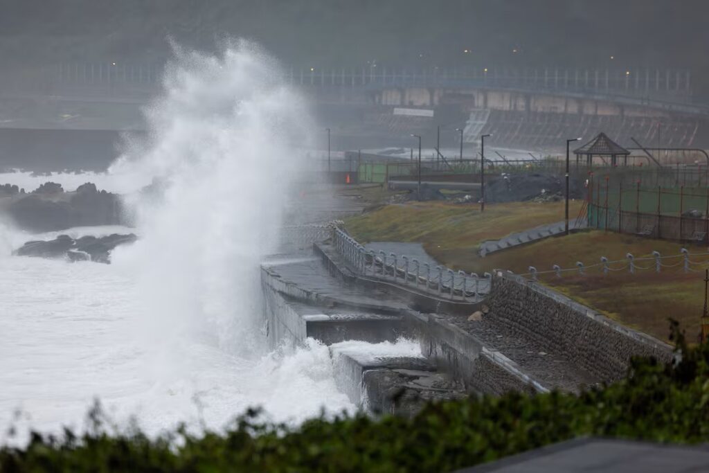 Ondas fortes quebram contra uma estrutura de proteção na costa, com fumaça e neblina ao fundo e um estádio ao longe, em cenário de forte clima marítimo.