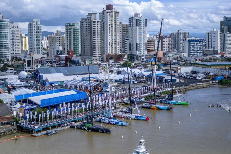 Vista aérea de um porto fluvial em cidade brasileira, com barcos e veleiros ao longo do canal e tendas montadas para um evento, ao lado de prédios altos